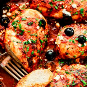 Tender cooked chicken breasts simmer in a vibrant tomato sauce, garnished with chopped parsley, sliced black olives, and crumbled feta cheese—all beautifully arranged in a skillet with a fork holding one succulent piece. Above the dish, “Mediterranean Chicken Recipe” is elegantly displayed.
