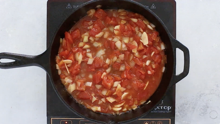 Top-down view of chopped tomatoes and onions simmering in a black cast iron skillet placed on an induction cooktop, forming the flavorful base for a Mediterranean Chicken Skillet Recipe.