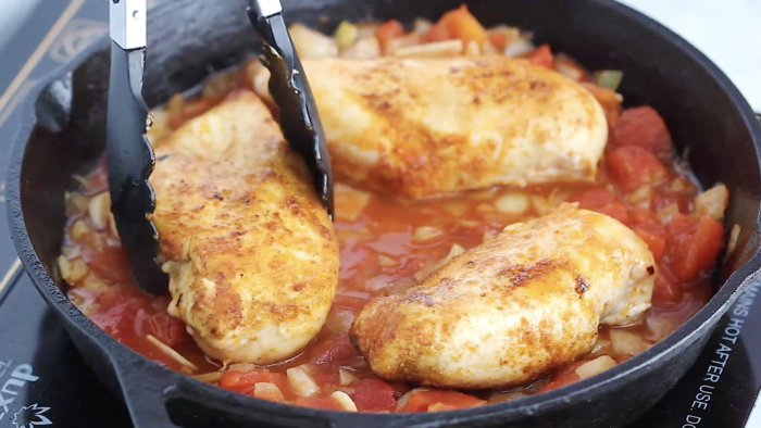 Chicken breasts being cooked in a cast iron skillet with a chunky tomato and vegetable sauce create a flavorful Mediterranean Chicken Skillet Recipe; metal tongs are holding one piece of chicken.