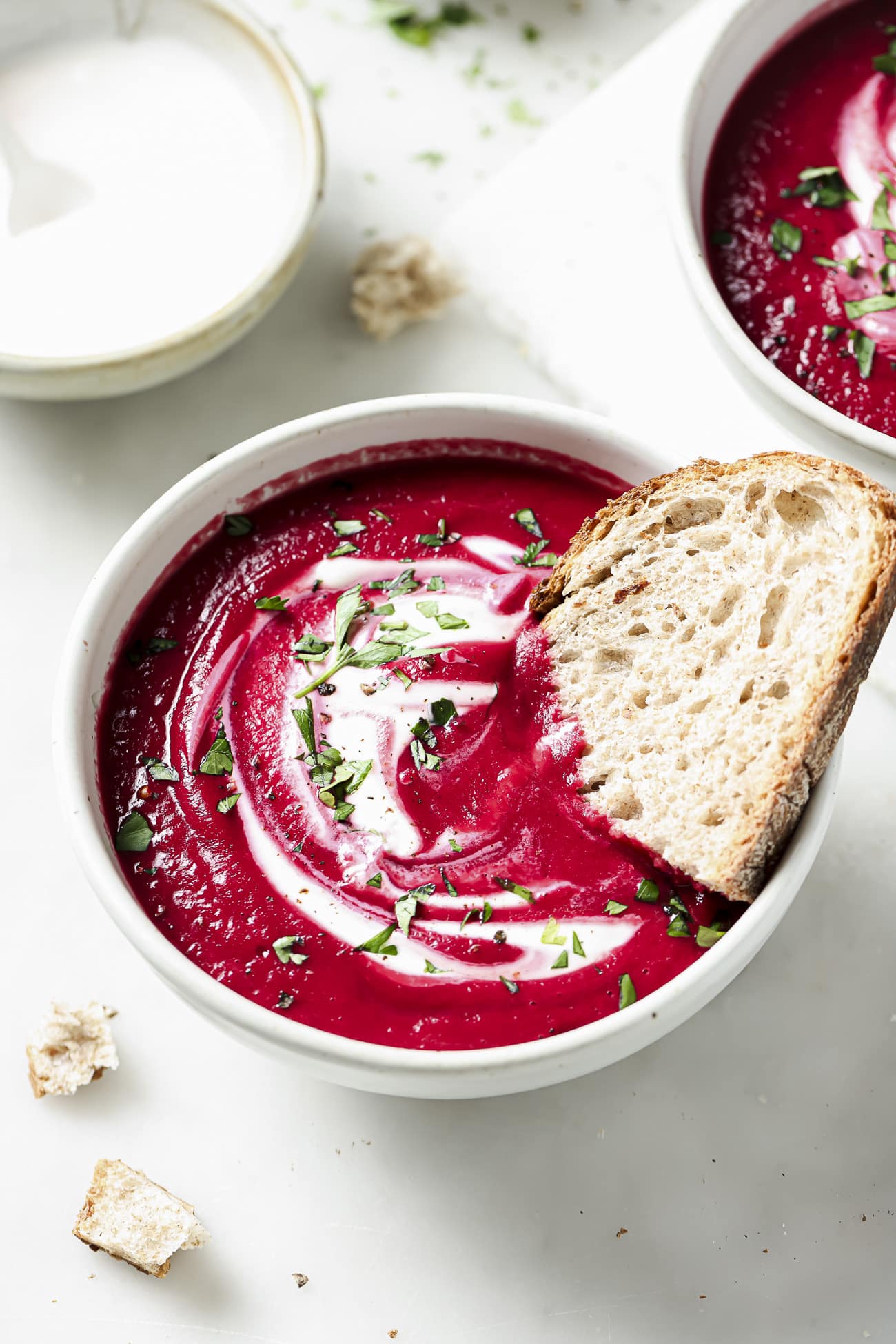 Beet soup in a white bowl with garnish and a slice of bread.