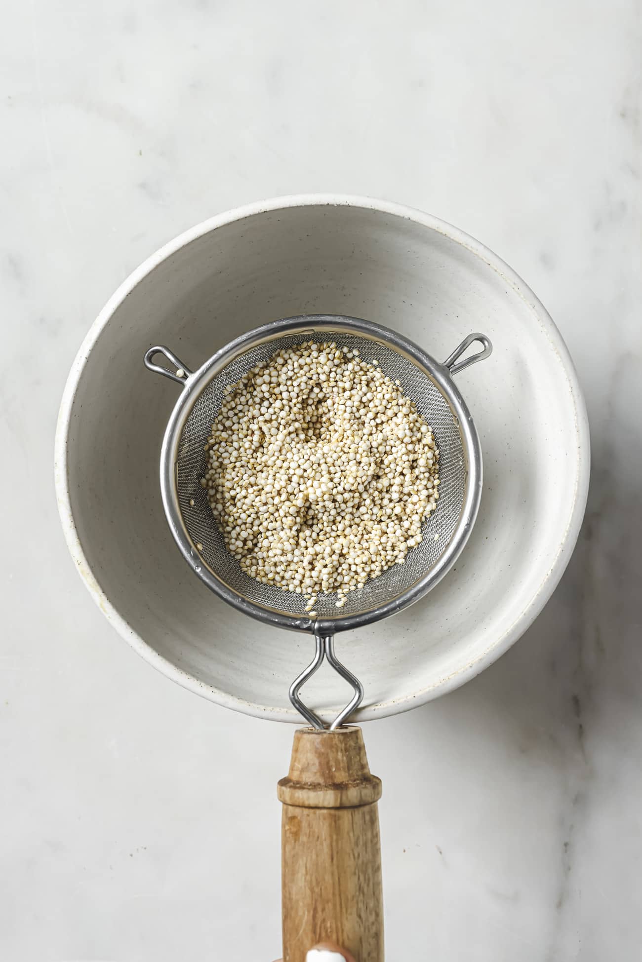 Uncooked quinoa in a fine mesh strainer over a white bowl. 