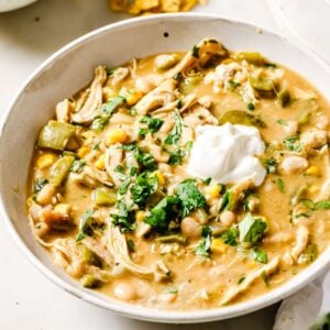 A bowl of creamy White Chicken Chili topped with a dollop of sour cream and chopped fresh cilantro, surrounded by small bowls of cilantro and crispy tortilla chips on a white surface.