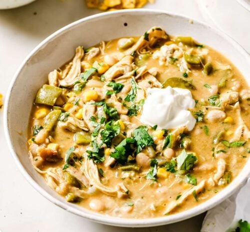 A bowl of creamy White Chicken Chili topped with a dollop of sour cream and chopped fresh cilantro, surrounded by small bowls of cilantro and crispy tortilla chips on a white surface.