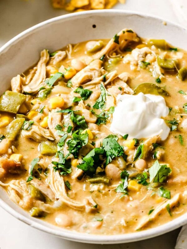 A bowl of creamy White Chicken Chili topped with a dollop of sour cream and chopped fresh cilantro, surrounded by small bowls of cilantro and crispy tortilla chips on a white surface.