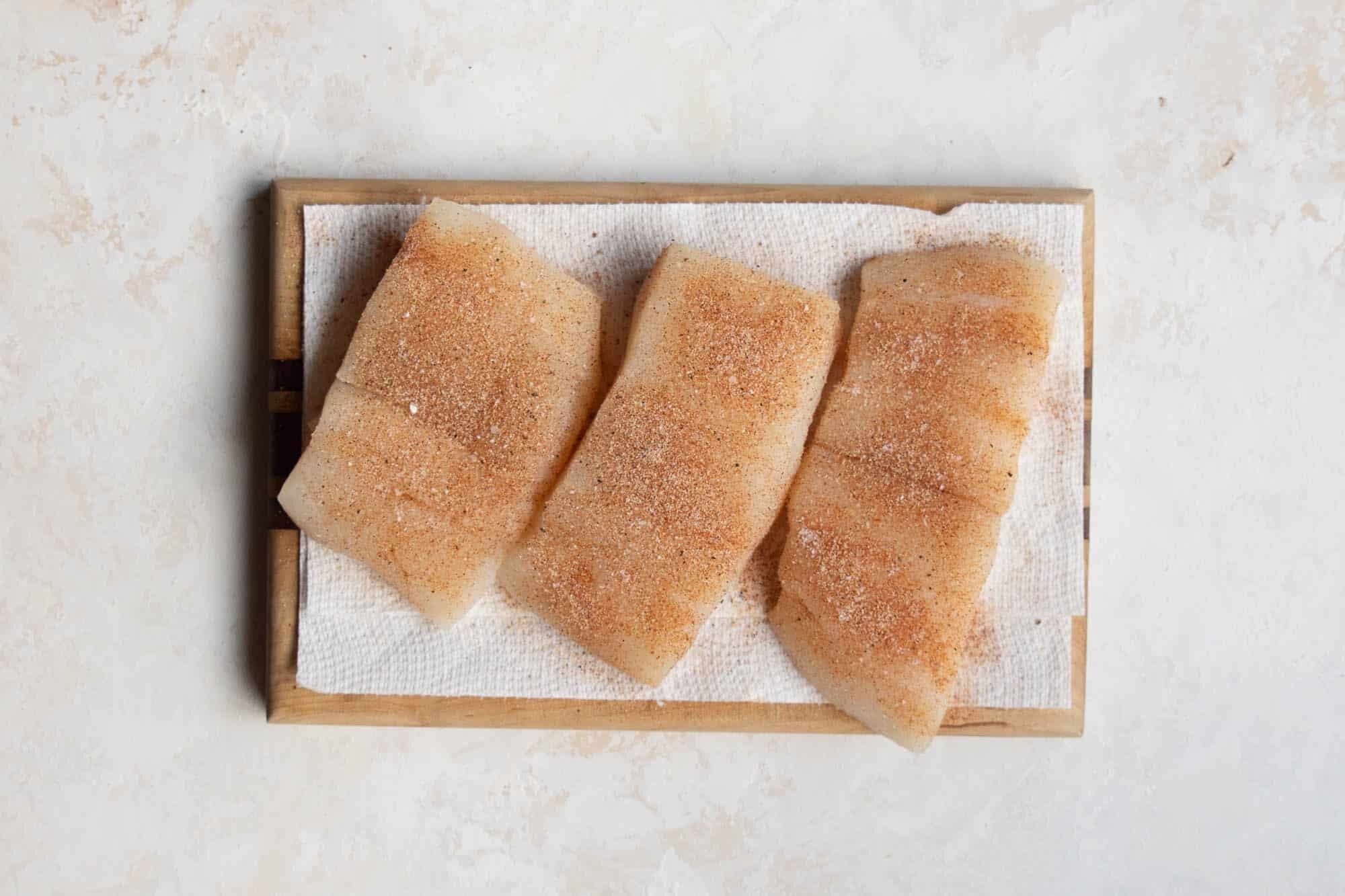 Three pieces of raw halibut fillets, scored and sprinkled with seasoning, rest on a paper towel on a wooden cutting board&mdash;a perfect start to your next halibut recipe.