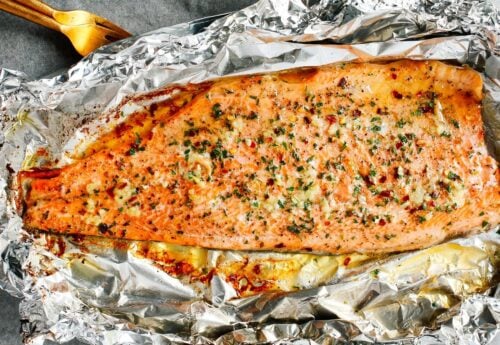 A large, seasoned baked salmon fillet sits on crinkled aluminum foil, garnished with herbs and spices, with a golden, slightly crispy surface. A serving spatula is visible in the corner.