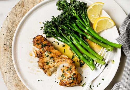 Air Fryer Cod fillet with steamed broccolini and lemon wedges on a white plate, garnished with chopped herbs. A small bowl of herbs, a plate with lemon slices, and a fork rest nearby on a marble surface.