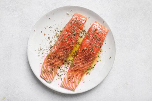 Two raw salmon fillets on a white plate, drizzled with olive oil and sprinkled with dried herbs, ready to be transformed into delicious oven baked salmon on a light-colored surface.