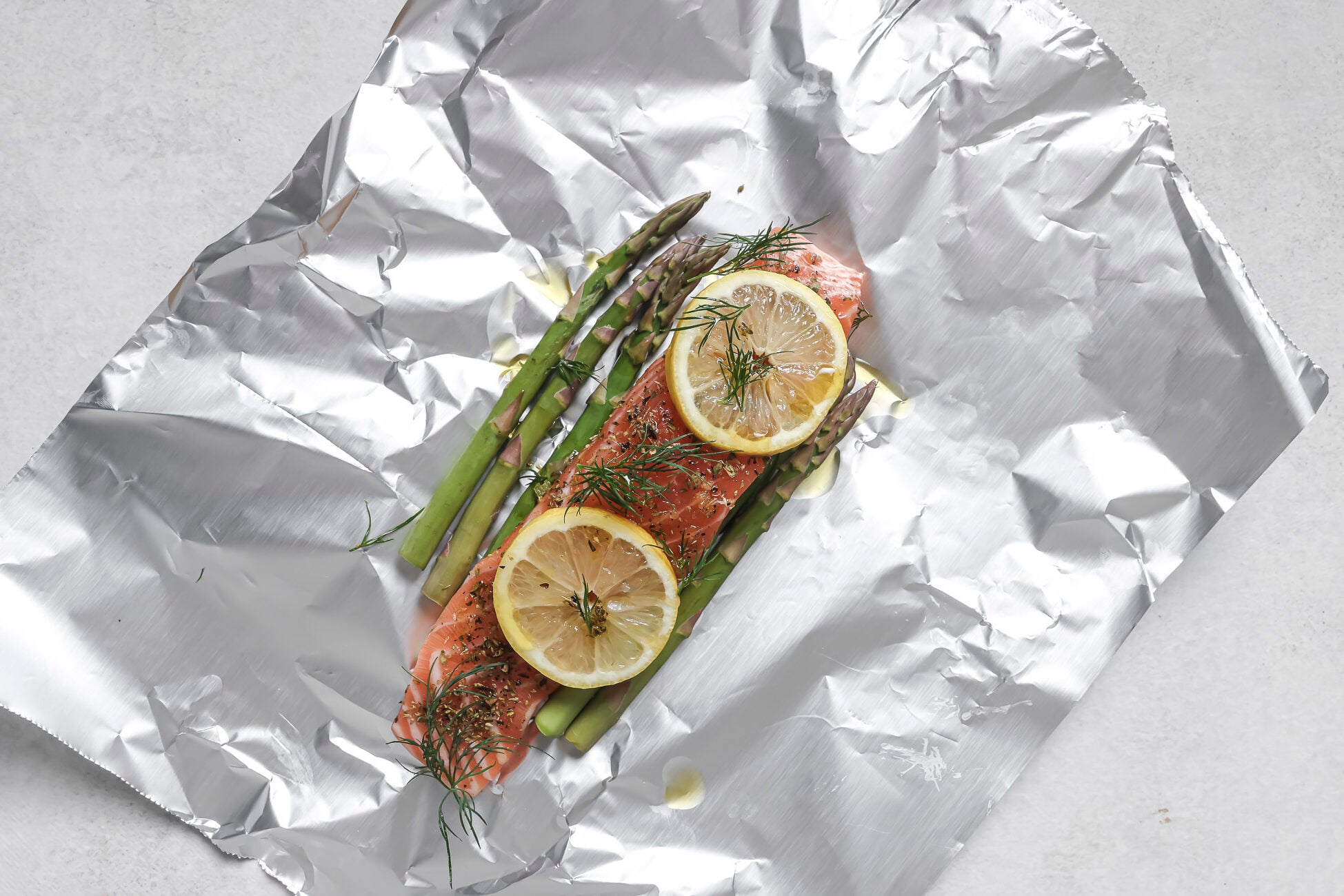 Salmon fillet topped with lemon slices, fresh dill, and asparagus spears on a sheet of aluminum foil, ready to be wrapped and cooked.