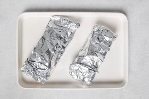 Two oven baked salmon fillets wrapped in aluminum foil are placed side by side on a rectangular white tray against a light gray background.