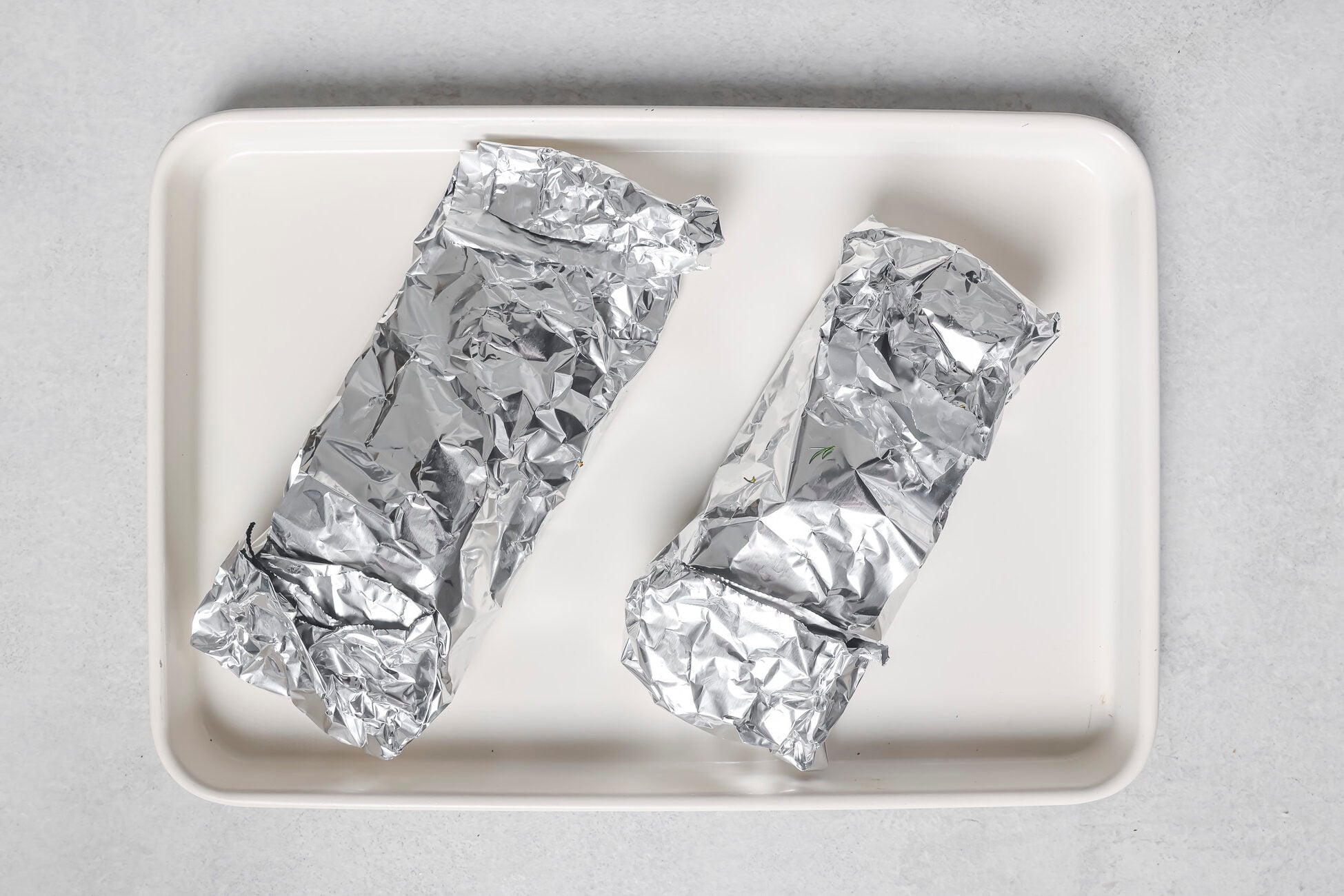 Two salmon fillets wrapped in aluminum foil are placed side by side on a rectangular white tray against a light gray background.
