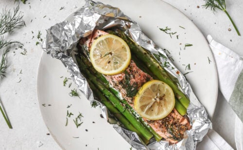 Two servings of oven baked salmon with asparagus and lemon slices in foil packets, garnished with fresh dill, served on white plates. Spices, herbs, and lemon wedges are artfully arranged around the plates on a light surface.