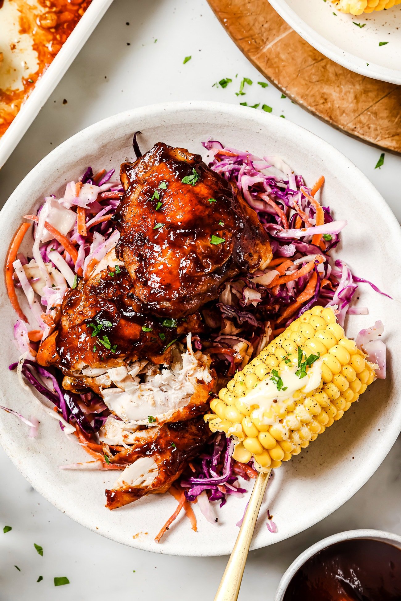 Baked BBQ chicken thighs on a plate with coleslaw and corn.