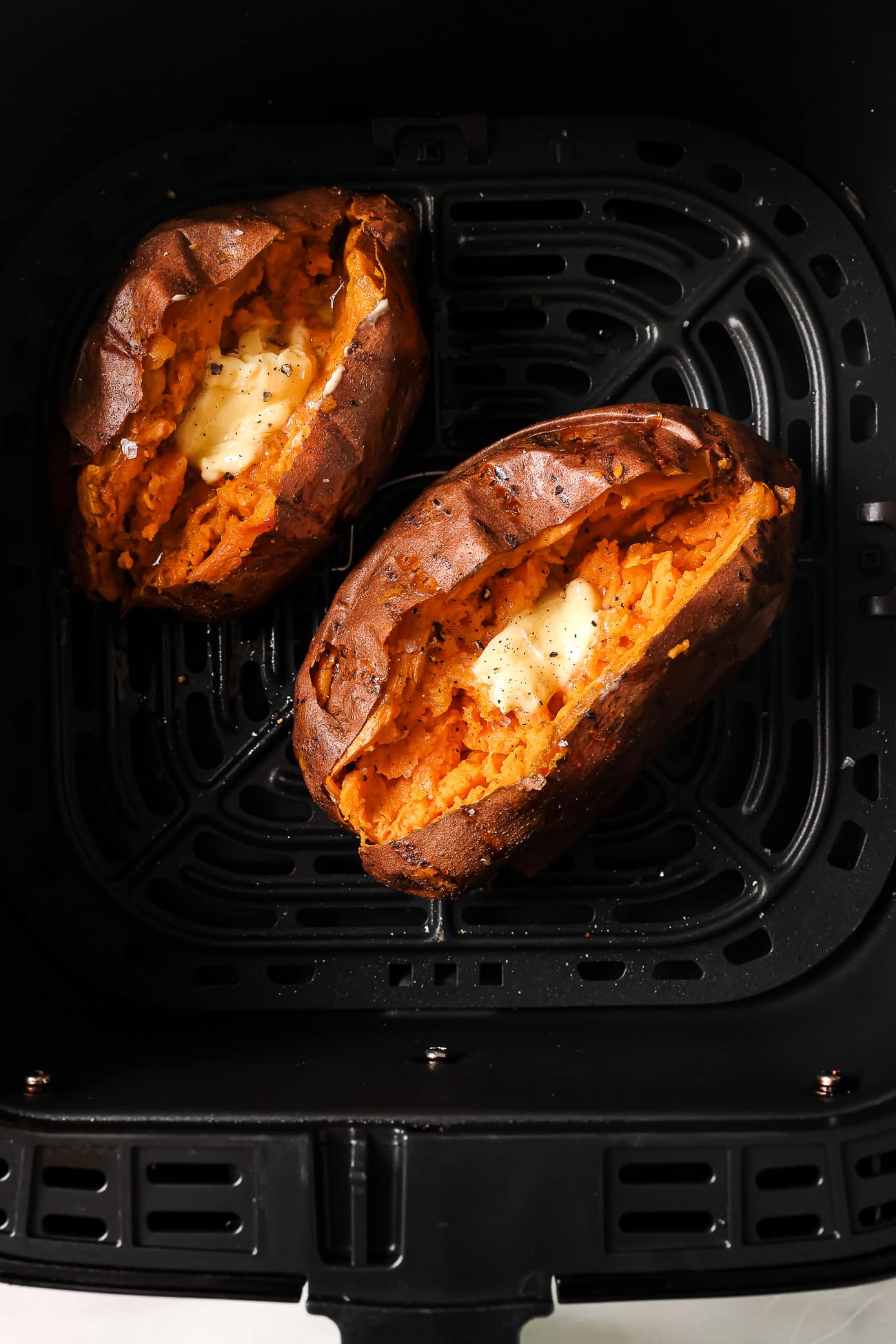 Two sweet potatoes cut open and topped with butter in an air fryer basket.