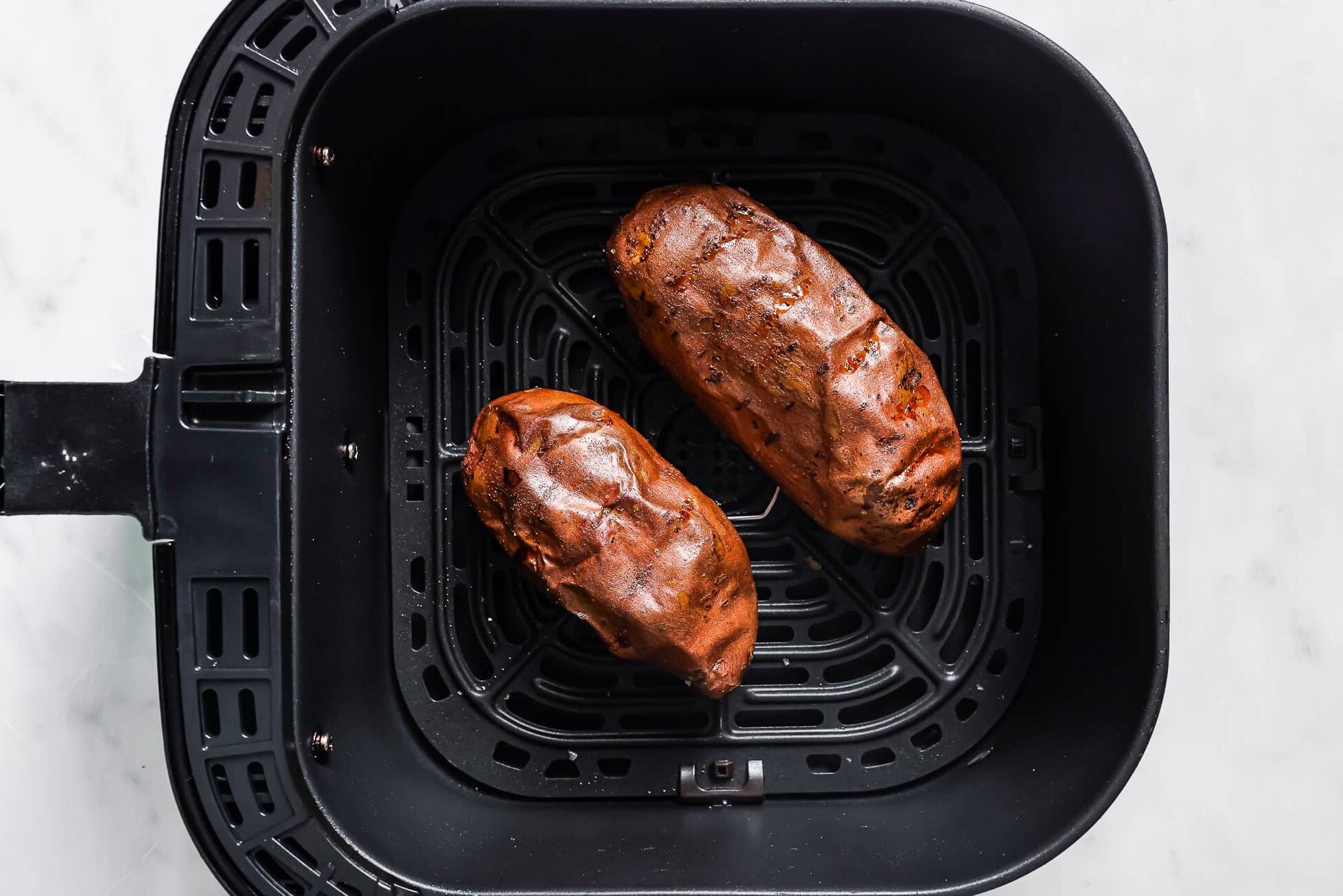 Two cooked Air Fryer Baked Sweet Potatoes sit inside the basket of an air fryer on a white marble surface.