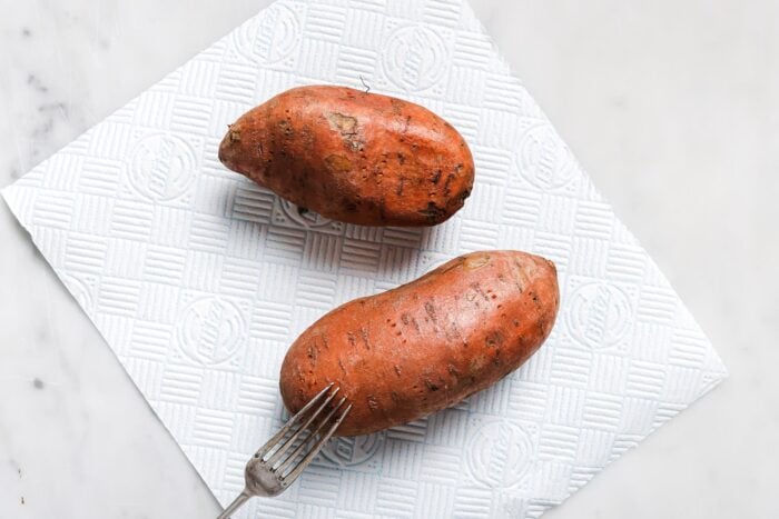 Two whole Air Fryer Baked Sweet Potatoes rest on a white textured paper towel on a marble surface, with a fork placed beside them.
