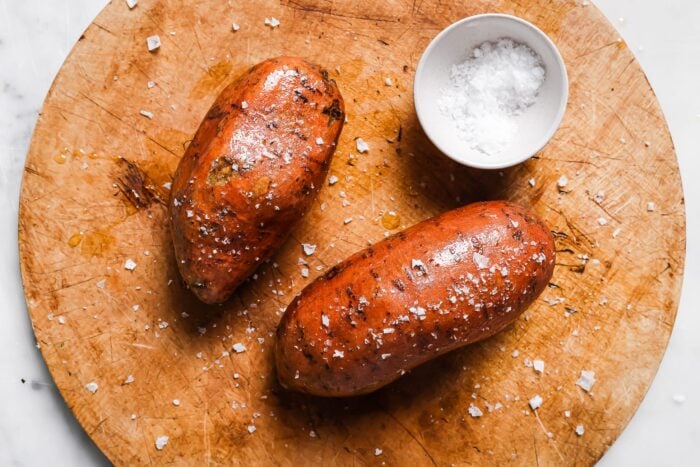 Two Air Fryer Baked Sweet Potatoes rest on a round wooden cutting board, sprinkled with coarse sea salt. A small white bowl filled with sea salt sits beside the sweet potatoes.