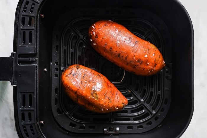 Two whole sweet potatoes with seasoned skin sit inside the basket, ready to become perfectly tender Air Fryer Baked Sweet Potatoes.