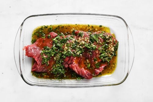 Raw steak in a glass dish sits in marinade with chopped herbs, minced garlic, and oil on a white background—perfectly prepared for making Carne Asada In The Oven.