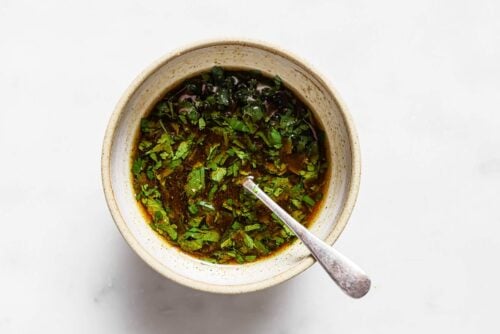 A bowl of chimichurri sauce with chopped herbs and oil, with a spoon resting inside, on a white surface—perfect for drizzling over Carne Asada In The Oven.