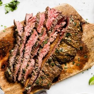 Sliced grilled steak on a wooden cutting board, garnished with herbs, showcases the savory flavors of Carne Asada In The Oven. A bowl of guacamole, a lime wedge, and cilantro are nearby on a white surface.