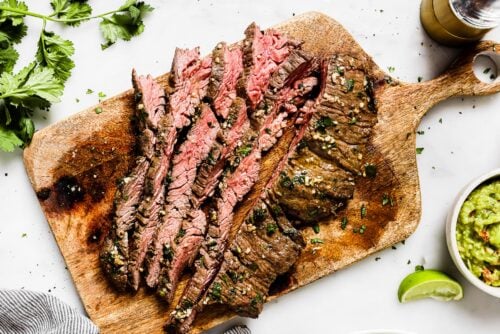 Sliced grilled steak on a wooden cutting board, garnished with herbs, showcases the savory flavors of Carne Asada In The Oven. A bowl of guacamole, a lime wedge, and cilantro are nearby on a white surface.