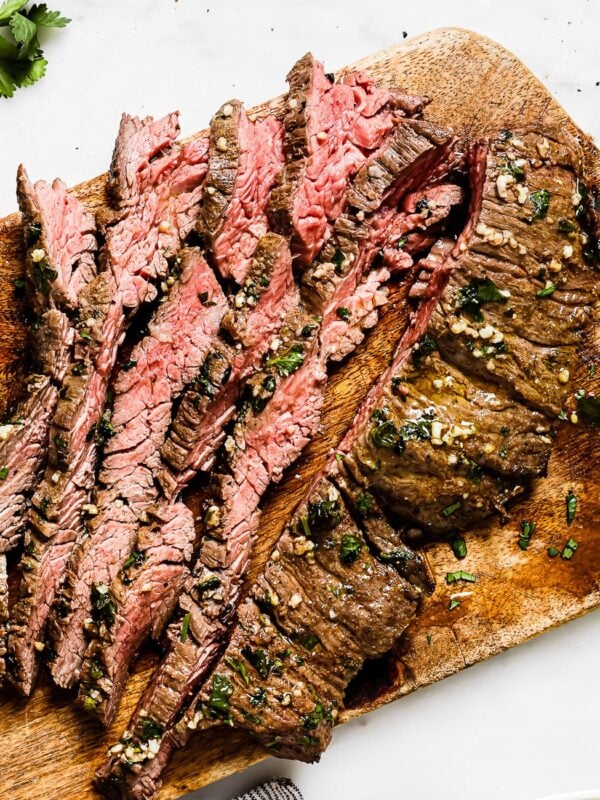 Sliced grilled steak on a wooden cutting board, garnished with herbs, showcases the savory flavors of Carne Asada In The Oven. A bowl of guacamole, a lime wedge, and cilantro are nearby on a white surface.