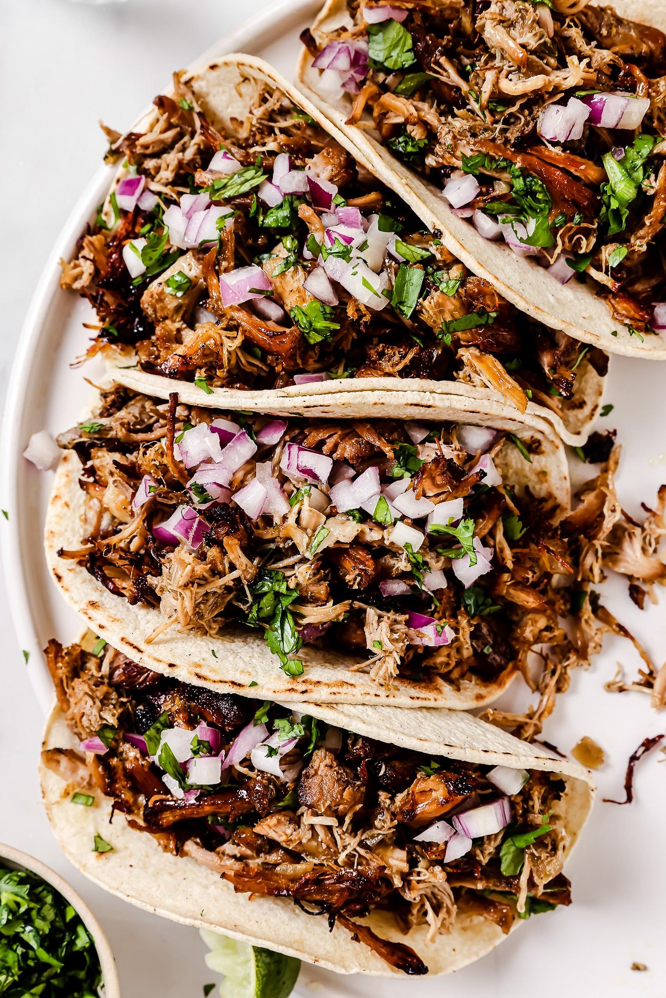 An overhead view of four carnitas tacos with toppings on a white plate. 