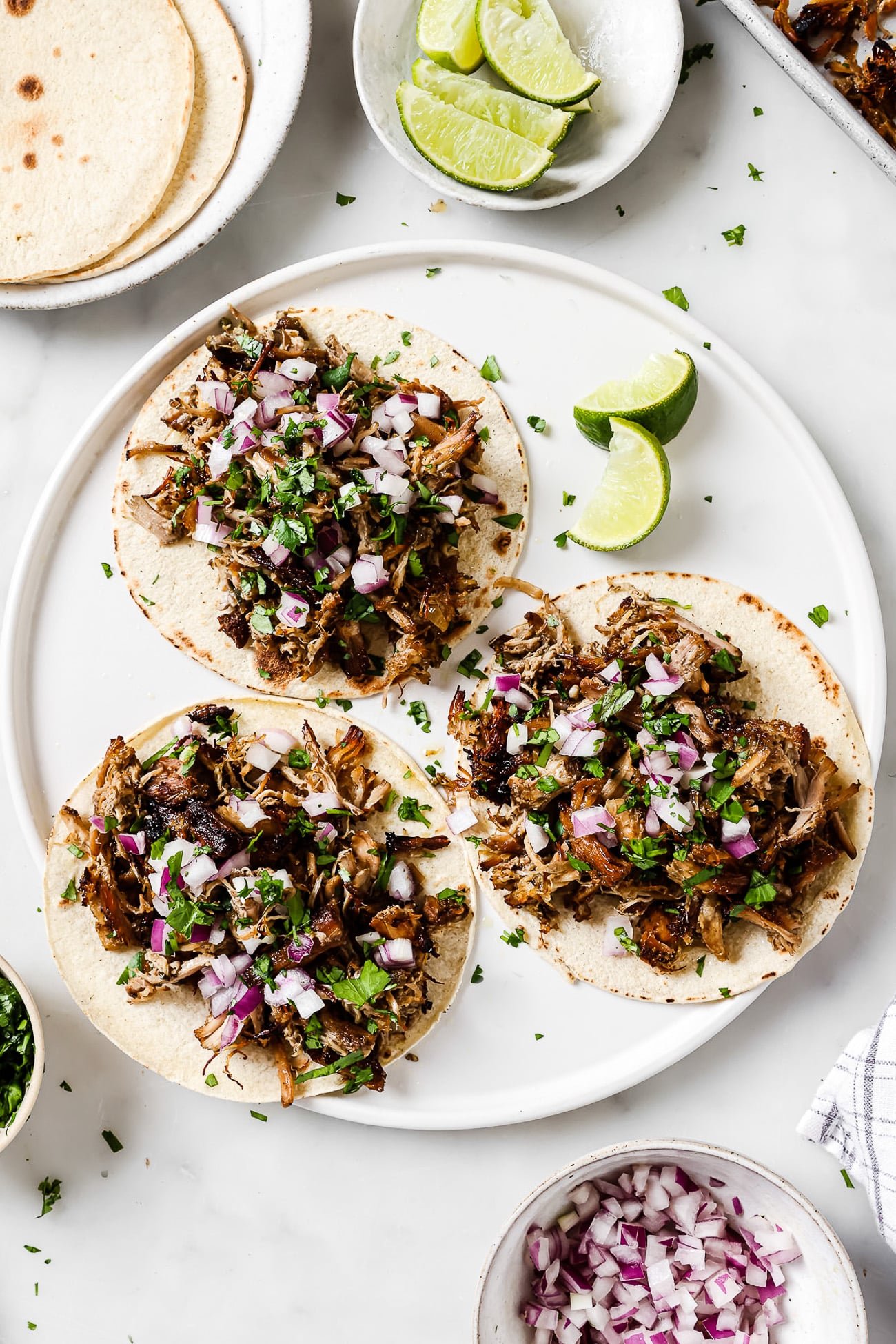 An overhead view of three carnitas tacos on a white plate with lime wedges on the side.