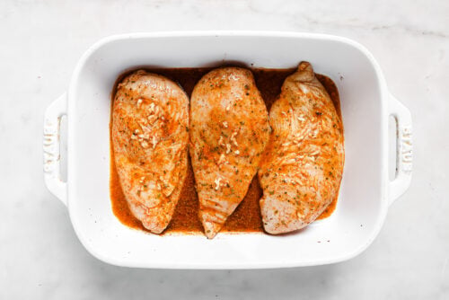 Three raw, seasoned chicken breasts marinating for Lemon Rosemary Chicken in a reddish liquid inside a white rectangular baking dish on a white marble surface.