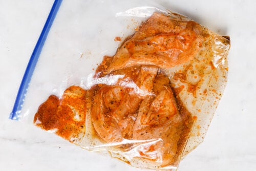 Lemon Rosemary Chicken pieces marinate in a clear resealable plastic bag with a reddish-brown marinade on a white surface.