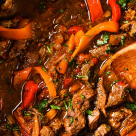 Close-up of Slow Cooker Pepper Steak with tender beef strips, red and yellow bell peppers, and a rich, savory sauce, garnished with chopped herbs and served in a black slow cooker with a wooden spoon.