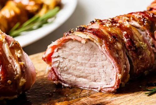 Sliced Bacon Wrapped Pork Tenderloin rests on a wooden cutting board, surrounded by herbs, with a plate of roasted potatoes softly blurred in the background.