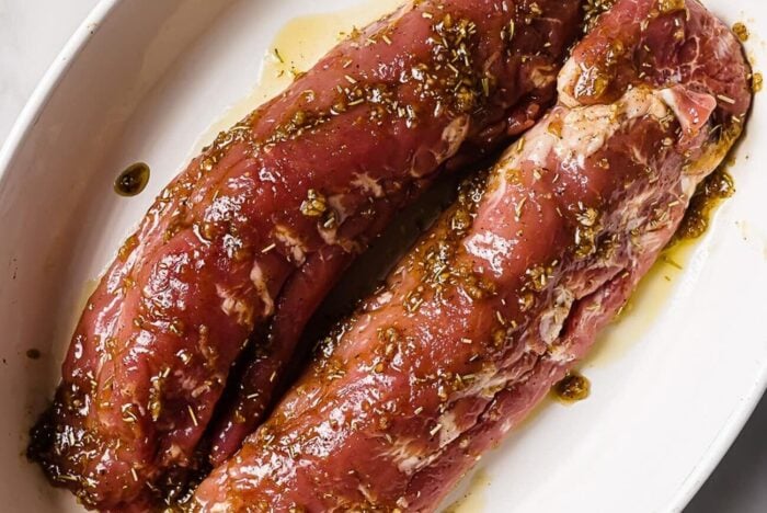 Two raw pork tenderloins, perfect for Bacon Wrapped Pork Tenderloin, are coated in a marinade with herbs and oil, placed side by side in a white oval dish.