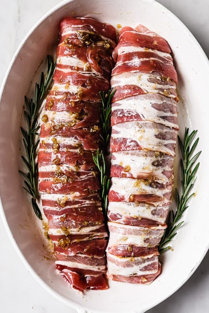 Two bacon wrapped pork tenderloins in a white baking dish with rosemary sprigs around it. 