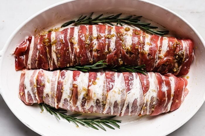 Two Bacon Wrapped Pork Tenderloin pieces rest in a white baking dish, garnished with fresh rosemary sprigs and drizzled with a savory marinade or seasoning.