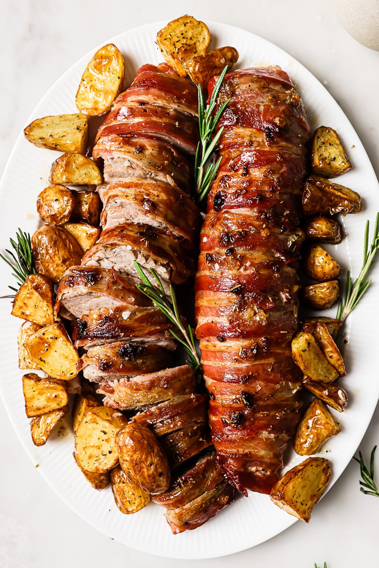 Sliced bacon wrapped pork tenderloin on a serving platter with roasted potatoes. 