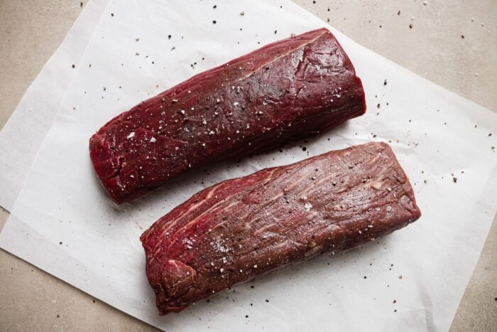 Two raw Garlic Butter Beef Tenderloin steaks on a sheet of white parchment paper, sprinkled with coarse salt and cracked black pepper, rest on a light-colored surface.