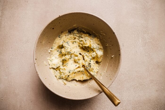 A beige ceramic bowl filled with herbed garlic butter mixture, ideal for pairing with Garlic Butter Beef Tenderloin, and a gold spoon resting inside, placed on a neutral, textured surface.
