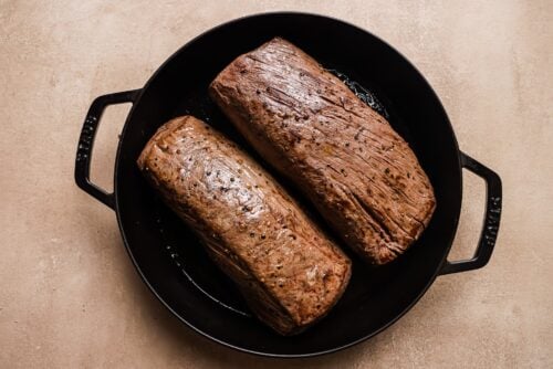 Two Garlic Butter Beef Tenderloin roasts seared in a black cast iron pan, resting on a light beige surface.