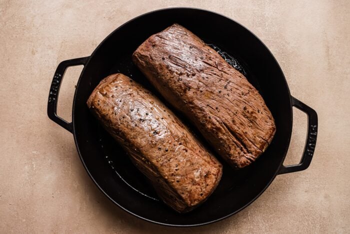 Two Garlic Butter Beef Tenderloin roasts seared in a black cast iron pan, resting on a light beige surface.