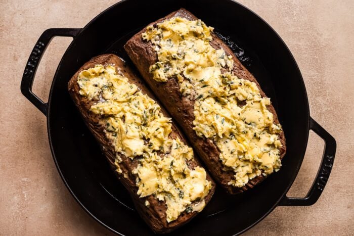 Two halved roasted eggplants in a black cast iron pan, topped with a generous layer of herbed butter—reminiscent of the richness found in Garlic Butter Beef Tenderloin—rest on a light brown surface.