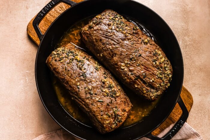 Two cooked Garlic Butter Beef Tenderloin roasts with a herb and garlic crust sit in a black cast iron pan with handles, surrounded by savory juices on a beige surface.