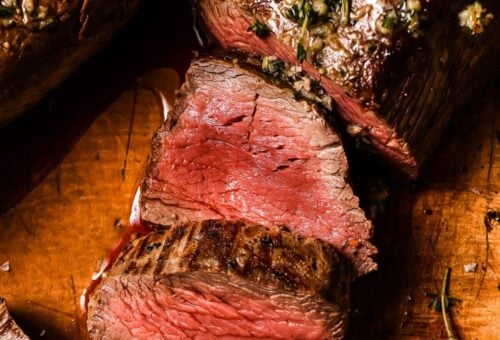Garlic Butter Beef Tenderloin sliced medium-rare on a wooden cutting board, topped with a savory garlic and herb mixture. The beef is juicy and pink inside with a beautifully browned exterior.