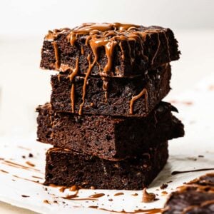 A stack of four pumpkin brownies drizzled with melted chocolate sits on white parchment paper, with crumbs scattered around.