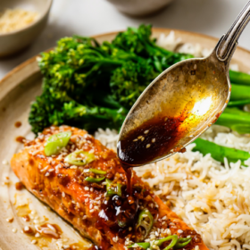 A plate with Honey Garlic Salmon fillet, white rice, and steamed broccoli. A spoon drizzles sauce over the salmon. Small bowls of sauce and green onions are in the background. Text reads Honey Garlic Salmon Recipe.