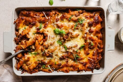 A Baked Ziti with Ground Beef casserole topped with melted cheese, fresh basil, and tomato sauce sits in a rectangular baking dish with a serving spoon, ready to be served.