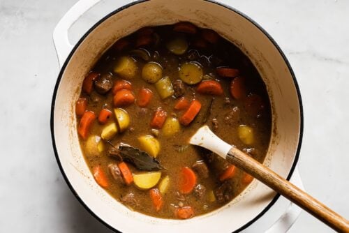 A white pot filled with Apple Cider Beef Stew—chunks of carrots, potatoes, and beef in a rich brown broth, accented by a bay leaf and a wooden spoon resting inside.