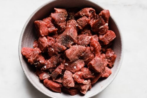 A bowl filled with raw, cubed beef pieces seasoned with salt and black pepper, ready to be used in an Apple Cider Beef Stew, placed on a white surface.