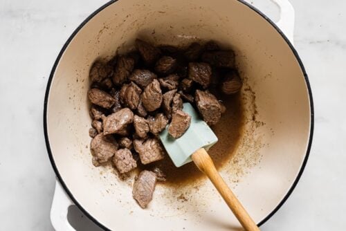 Chunks of browned beef for Apple Cider Beef Stew sit in a large white pot with some liquid at the bottom. A light blue spatula with a wooden handle rests in the pot, stirring the meat. The pot is on a white surface.